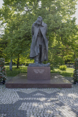 Zdjęcie wykonane na wprost, przedstawiające całopostaciową, rzeźbę mężczyzny w podeszłym wieku. Pomnik ustawiony jest na niskim, prostopadłościennym postumencie wykonanym z czerwonego granitu, na którego ścianie bocznej zostały wyryte litery układające się w napis: „Marszałek Polski, Józef Piłsudski, 1867-1935”. Figura mężczyzny ubrana jest w mundur oraz długi płaszcz. Mężczyzna jest lekko pochylony przed siebie z poważnym wyrazem twarzy, natomiast dłonie ma splecione za plecami. Mężczyzna ma krótkie, zaczesane do tyłu włosy oraz gęste brwi i długie, zakrywające praktycznie całe usta wąsy.