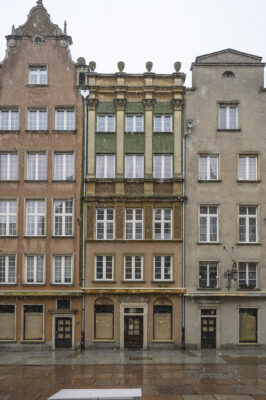 Zdjęcie pionowe, wykonane na wprost, przedstawiające trzy, pięciokondygnacyjne kamienice w tkance zabytkowej Głównego Miasta w Gdańsku. Ich fasady utrzymane są w odcieniach jasnych brązów, zieleni oraz ciemnych beży. Każda z kamienic posiada różnie zdobiony szczyt.