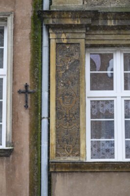 Zdjęcie pionowe przedstawiające zbliżenie na detal dekoracyjny znajdujący się na brązowej fasadzie jednej z kamienic w tkance zabytkowej Głównego Miasta w Gdańsku. Dekoracja została wykonana w technice sgraffito i składa się na nią ornament okuciowy z girlandami. W centralnym punkcie ornamentu umieszczono popiersie uśmiechniętego człowieka w kapeluszu.