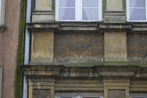 Zdjęcie poziome przedstawiające zbliżenie na detal dekoracyjny znajdujący się na brązowej fasadzie jednej z kamienic w tkance zabytkowej Głównego Miasta w Gdańsku. Dekoracja została wykonana w technice sgraffito i składa się na nią ornament okuciowy z girlandami.