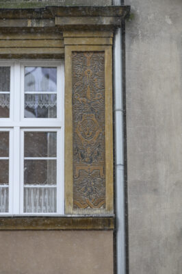 Zdjęcie pionowe przedstawiające zbliżenie na detal dekoracyjny znajdujący się na brązowej fasadzie jednej z kamienic w tkance zabytkowej Głównego Miasta w Gdańsku. Dekoracja została wykonana w technice sgraffito i składa się na nią ornament okuciowy z girlandami. W centralnym punkcie ornamentu umieszczono popiersie uśmiechniętego człowieka w czepcu.