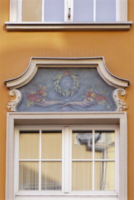 Zdjęcie pionowe ze zbliżeniem na okno pomarańczowej kamienicy, nad którym znajduje się malarska dekoracja architektoniczna. W kwaterze o nieregularnym kształcie umieszczono malowidło przedstawiające dwa rogi obfitości wypełnione owocami oraz roślinnością. Są one rozłożone względem siebie symetrycznie, zaś nad nimi, na osi pionowej znajduje się zielony wieniec laurowy z czerwoną wstęgą.