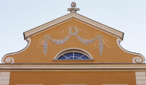 Zdjęcie poziome ze zbliżeniem na szczyt pomarańczowej kamienicy. Ozdobiony jest on malowidłem przedstawiającym roślinną girlandę, bogato ozdobioną owocami. Rozłożona jest ona symetrycznie wzdłuż osi pionowej, zaś podtrzymują ją trzy wieńce laurowe – dwa mniejsze znajdujące się po bokach oraz większy umieszczony u szczytu kompozycji.