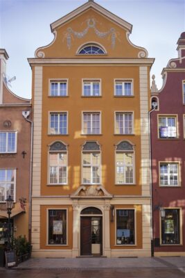 Zdjęcie pionowe przedstawiające pięciokondygnacyjną kamienicę, która znajduje się w zabytkowej tkance Gdańska. Jej pomarańczowa fasada ozdobiona jest boniowaniem, zaś nad wejście znajduje się klasycyzujący pseudo portal.