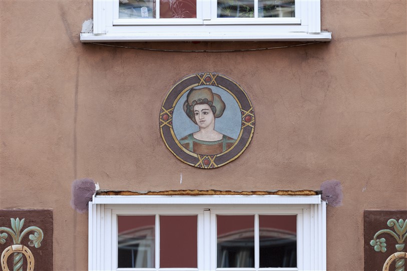 Zdjęcie poziome ze zbliżeniem na dekorację architektoniczną, która znajduje się pomiędzy dwoma oknami pomarańczowo-brązowej kamienicy. W okrągłym medalionie umieszczono malowidło przedstawiające popiersie kobiety w historyzującym stroju.