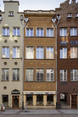 Zdjęcie pionowe przedstawiające czterokondygnacyjną kamienicę, która znajduje się w zabytkowej tkance Gdańska. Jej pomarańczowa fasada jest bogato zdobiona brązowym ściennym sgraffito. Wzdłuż jej ścian bocznych znajdują się szare, długie rynny deszczowe, które wizualnie oddzielają budynek od sąsiadujących z nim kamienic.