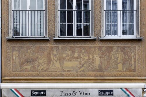 Zdjęcie poziome ze zbliżeniem na malarską dekorację architektoniczną, która znajduje się na fasadzie pomarańczowej kamienicy. Jest to brązowe, szczegółowo wykonane, sgraffito ścienne. Przedstawia ono sceny rodzajowe z życia gdańskich patrycjuszy w historycznych strojach.