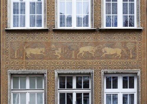 Zdjęcie poziome ze zbliżeniem na malarską dekorację architektoniczną, która znajduje się na fasadzie pomarańczowej kamienicy. Jest to brązowe, szczegółowo wykonane, sgraffito ścienne. Przedstawia ono scenę animalistyczną, w której wzajemnie walczą ze sobą dwa jednorożce z lwem oraz jeleniem.