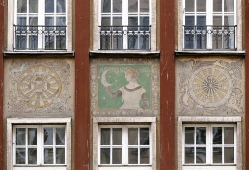 Zdjęcie poziome ze zbliżeniem na malarską dekorację architektoniczną, która znajduje się na fasadzie ciemnoczerwonej kamienicy. Są to trzy, wielobarwne sgraffita ścienne umieszczone w kwadratowych kwaterach między oknami budynku. Środkowe przedstawia portret kobiety w historyzującym stroju na zielonym tle. W uniesionej prawej dłoni trzyma ona księżyc w formie sierpa, zaś za jej plecami znajduje się napis: „Astronomia”. W bocznych kwaterach umieszczono bogato zdobione mapy nieboskłonu.
