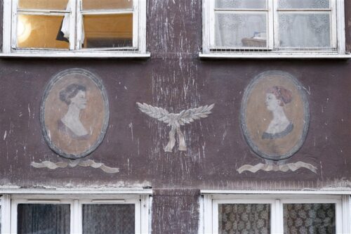 Zdjęcie poziome ze zbliżeniem na malarską dekorację architektoniczną, która znajduje się na fasadzie bordowo-brązowej kamienicy. Jest to wielobarwne sgraffito, które umieszczono na dwóch owalnych medalionach. Przedstawia ono dwa, profilowe portrety kobiece w historycznych strojach. Postaci zwrócone są ku sobie, zaś pomiędzy znajduje się jasnobeżowy ornament rośliny.