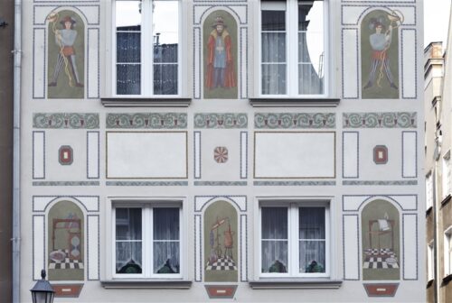 Zdjęcie poziome ze zbliżeniem na malarską dekorację architektoniczną, która znajduje się na fasadzie jasnoszarej kamienicy. Jest to wielobarwne, szczegółowo wykonane sgraffito ścienne. Przedstawia ono wizerunki trzech uczonych w historycznych strojach umieszczonych w półokrągło zamkniętych kwaterach. Poniżej znajdują się identyczne kwatery jednak przedstawiają one różnego rodzaju narzędzia badawcze.