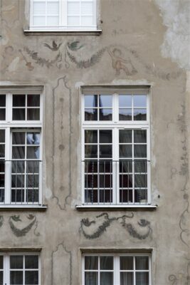 Zdjęcie pionowe ze zbliżeniem na malarską dekorację architektoniczną, która znajduje się na fasadzie jasnobeżowej kamienicy. Jest to wielobarwne sgraffito ścienne, które zostało umieszczone bezpośrednio na ścianie budynku. Przedstawia ono unoszące się putta z instrumentami muzycznymi, a także girlandy roślinne i symbolicznie wykonaną arabeskę.
