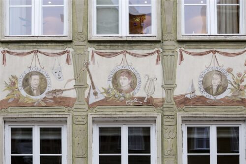 Zdjęcie pionowe ze zbliżeniem na malarską dekorację architektoniczną, która znajduje się na fasadzie ciemnozielonej kamienicy. Jest to wielobarwna dekoracja ścienna przedstawiająca trzy okrągłe medaliony z portretami artystów: Alojzego Żółkowskiego, Heleny Modrzejewskiej i Wojciecha Bogusławskiego. Dodatkowo, każdy z medalionów ozdobiony jest różnymi instrumentami muzycznymi i przedmiotami teatralnymi.