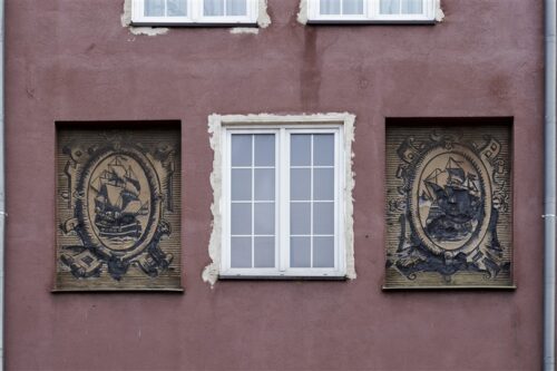 Zdjęcie poziome ze zbliżeniem na malarską dekorację architektoniczną, która znajduje się na fasadzie bordowej kamienicy. Dwie beżowe, kwadratowe kwatery zostały ozdobione ciemnym sgraffito ściennym o motywie marynistycznym. Całość przypomina dwie nowożytne grafiki wykonane w technice miedziorytu.