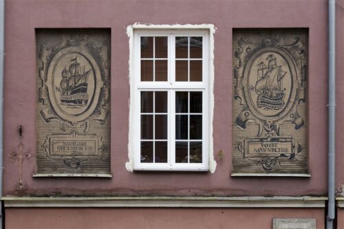 Zdjęcie poziome ze zbliżeniem na malarską dekorację architektoniczną, która znajduje się na fasadzie bordowej kamienicy. Dwie beżowe, kwadratowe kwatery zostały ozdobione ciemnym sgraffito ściennym o motywie marynistycznym. Całość przypomina dwie nowożytne grafiki wykonane w technice miedziorytu.