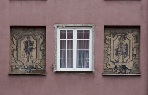 Zdjęcie poziome ze zbliżeniem na malarską dekorację architektoniczną, która znajduje się na fasadzie bordowej kamienicy. Dwie beżowe, kwadratowe kwatery zostały ozdobione ciemnym sgraffito ściennym z całopostaciowymi przedstawieniami mężczyzn w historycznych strojach. Całość przypomina dwie nowożytne grafiki wykonane w technice miedziorytu.