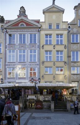 Zdjęcie pionowe przedstawiające trzy wielokolorowe kamienice w zabytkowej tkance Gdańska. Wzdłuż ich ścian bocznych znajdują się szare, długie rynny deszczowe, które wizualnie oddzielają budynki od sąsiadujących z nimi kamienic.