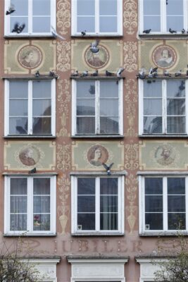 Zdjęcie pionowe ze zbliżeniem na malarską dekorację architektoniczną, która znajduje się na fasadzie pomarańczowej kamienicy. Przedstawia ona sześć, jasnozielonych, prostokątnych kwater z okrągłym medalionem w każdej. Zostały w nich umieszczone portrety artystów i pisarzy z różnych kręgów kulturowych Europy, np. Dante Alighieri, Jan Kochanowski czy William Shakespeare. Kwatery są ułożone horyzontalnie i znajdują się między oknami budynku, natomiast wertykalnie umieszczono jasnobeżowe, symetryczne arabeski oparte na osi pionowej.