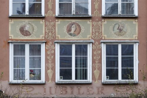 Zdjęcie poziome ze zbliżeniem na malarską dekorację architektoniczną, która znajduje się na fasadzie pomarańczowej kamienicy. Przedstawia ona trzy, jasnozielonych, prostokątnych kwater z okrągłym medalionem w każdej. Zostały w nich umieszczone portrety artystów i pisarzy z różnych kręgów kulturowych Europy: Donato Bramante, Rafael Santi i Donatello. Kwatery są ułożone horyzontalnie i znajdują się między oknami budynku, natomiast wertykalnie umieszczono jasnobeżowe, symetryczne arabeski oparte na osi pionowej.