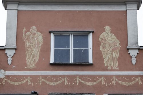 Zdjęcie poziome ze zbliżeniem na malarską dekorację architektoniczną, która znajduje się w szczycie pomarańczowej kamienicy. Jest to jasnożółte sgraffito ścienne, które przedstawia Platona i Arystotelesa. Wizerunki filozofów zostały zaczerpnięte ze Szkoły Astańskiej autorstwa Rafaela Santi.