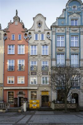 Zdjęcie pionowe przedstawiające trzy wielokolorowe, bogato zdobione kamienice w zabytkowej tkance Gdańska. Wzdłuż ich ścian bocznych znajdują się szare, długie rynny deszczowe, które wizualnie oddzielają budynki od sąsiadujących z nimi kamienic.