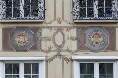 Zdjęcie poziome ze zbliżeniem na malarską dekorację architektoniczną, która znajduje się na fasadzie jasnobeżowej kamienicy. Przedstawia ona dwa, okrągłe jasnozielone medaliony otoczone obfitym wieńcem laurowym. Umieszczono na nich odrealnione, portretowe przedstawienia ludzkie. Medaliony znajdują się w ciemnozielonych kwaterach w formie oktagonu. Pomiędzy nimi umieszczono ornament roślinny układający się owalną formę.