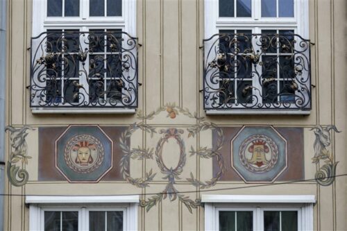 Zdjęcie poziome ze zbliżeniem na malarską dekorację architektoniczną, która znajduje się na fasadzie jasnobeżowej kamienicy. Przedstawia ona dwa, okrągłe jasnozielone medaliony otoczone obfitym wieńcem laurowym. Umieszczono na nich odrealnione, portretowe przedstawienia ludzkie. Medaliony znajdują się w ciemnozielonych kwaterach w formie oktagonu. Pomiędzy nimi umieszczono ornament roślinny układający się owalną formę, zaś po bokach są dwie, antropomorficzne głowy kozłów.