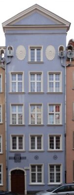 Zdjęcie pionowe ukazujące fasadę kamienicy w kolorze szaroniebieskim z elementami dekoracji metaloplastycznych i płaskorzeźbiarskich.Metaloplastyczne elementy zbudowane są z uproszczonych, zgeometryzowanych form floralnych odwołujących się do pierwotnej dekoracji – półokrągłych rozet kwiatowych wykonanych z kamienia, flankujących okno piątej kondygnacji.