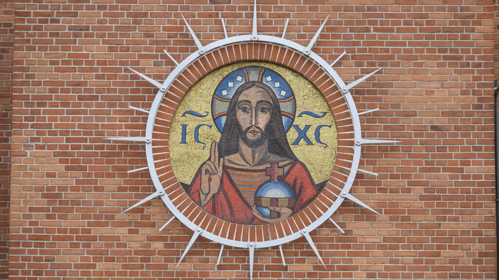 Zdjęcie poziome ze zbliżeniem na okrągłą, kolorową mozaikę z przedstawieniem Chrystusa Salvator Mundi – Chrystusa Zbawiciela Świata. Chrystus w czerwonej szacie umieszczony jest na złotym tle. Po obydwu stronach jego nimbu znajduje się napis: IX – XC, czyli christogram. Chrystus ma uniesioną prawą dłoń w geście błogosławieństwa, zaś w lewej trzyma jabłko królewskie.