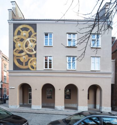 Zdjęcie przedstawia boczną elewację kamienicy z arkadowym podcieniem. Powyżej została umieszczona pionowa dekoracja wpisana w prostokąt, która przedstawia złote koła zębate zegarmistrzowskie na ciemnym tle.