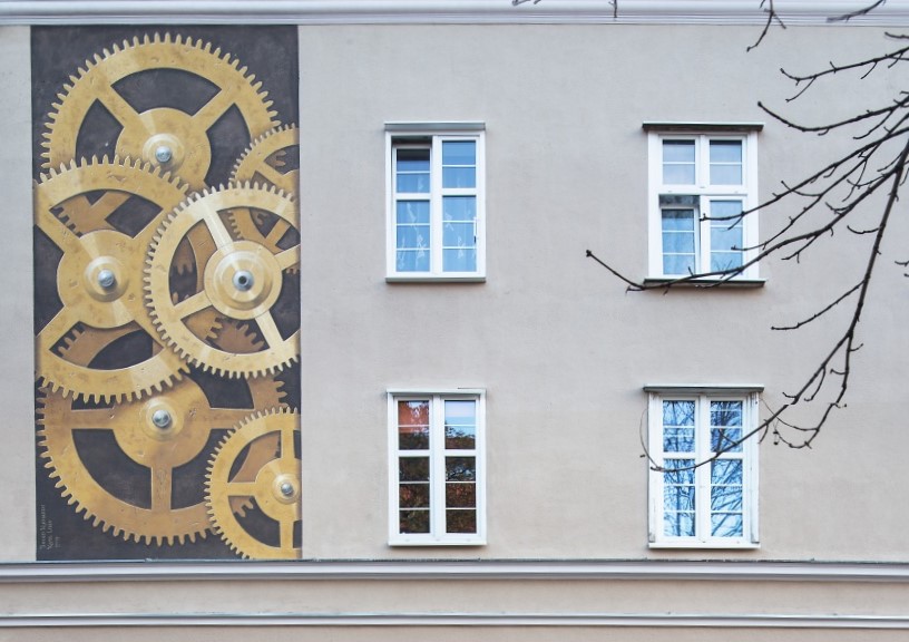 Zdjęcie przedstawia fragment bocznej elewacji kamienicy. Po lewej stronie, obok okien drugiej i trzeciej kondygnacji, została umiesczona pionowa dekoracja wpisana w prostokąt, która przedstawia złote koła zębate zegarmistrzowskie na ciemnym tle.