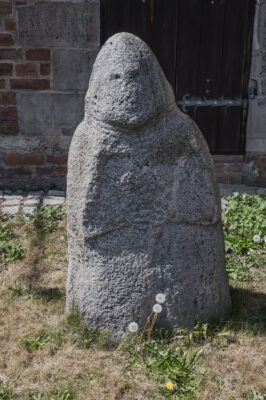 Zdjęcie pionowe przedstawiające niską rzeźbę kamienną o obłym kształcie. Ma ona stożkowaty kształt i dostrzegalne są cechy antropomorficzne. Na powierzchni rzeźby widoczne są zarysy rąk oraz dłoni, a także oczu, ust i nosa postaci.