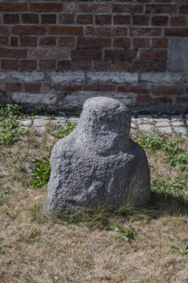 Zdjęcie pionowe przedstawiające niską rzeźbę kamienną o kształcie przypominającym ustawione na siebie dwa sześciany. Posiada ona antropomorficzne cechy. Na powierzchni rzeźby dostrzegalne są zarysy rąk, a także oczu, ust i nosa postaci.