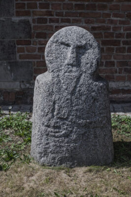 Zdjęcie pionowe przedstawiające niską rzeźbę kamienną o obłym kształcie, na który składa się sześcian i okrąg. Posiada ona antropomorficzne cechy. Na powierzchni rzeźby dostrzegalne są zarysy rąk oraz dłoni, a także oczu, ust i nosa postaci.