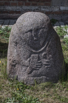 Zdjęcie pionowe przedstawiające niską rzeźbę kamienną o obłym kształcie. Ma ona stożkowaty kształt i dostrzegalne są cechy antropomorficzne. Na powierzchni rzeźby widoczne są zarysy rąk oraz dłoni, a także oczu, ust i nosa postaci.