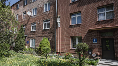 Zdjęcie poziome przedstawiające dwa ceglane budynki. W jednej ze ścian budynku po prawej stronie, znajduje się nisza, w której umieszczono kamienną rzeźbę. Przedstawia ona zgeometryzowaną postać stojącego mężczyzny. Przed wejściami do budynków posadzona została niska roślinność.