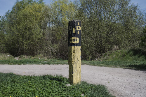 Zdjęcie poziome ukazujące w betonowy słup w formie prostopadłościanu, który jest ustawiony przy polnej drodze. Jego ściany zostały pomalowane jasnożółtą oraz czarną farbą. W efekcie słup przypomina czarną maskę umieszczoną na jasnożółtej podporze. Wygląd maski nasuwa skojarzenia z maskami rdzennych kultur afrykańskich lub południowoazjatyckich.