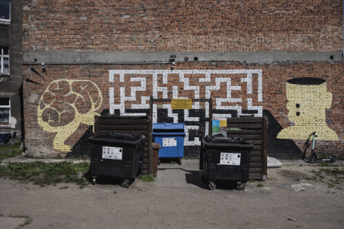 Zdjęcie poziome przedstawiające boczną ścianę ceglanego budynku, której powierzchnia została częściowo pokryta minimalistycznym graffiti. Przedstawia on schematycznie ukazany przekrój labiryntu. Po jego lewej stronie został ukazany w komiksowym stylu jasnożółty mózg, zaś po prawej przekrojona na wysokości czaszki głowa człowieka.