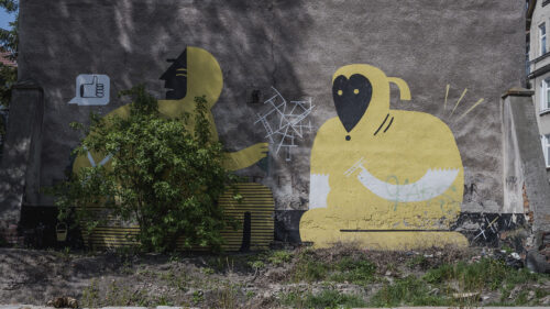 Zdjęcie poziome przedstawiające boczną ścianę szarego budynku, której powierzchnia została częściowo pokryta minimalistycznym graffiti z cyklu. Przedstawia on odrealnione dwie postaci ludzkie ukazane w komisowej estetyce. Postaci zostały wykonane jasnożółtą i białą farbą, która kontrastuje z ich czarnymi detalami.