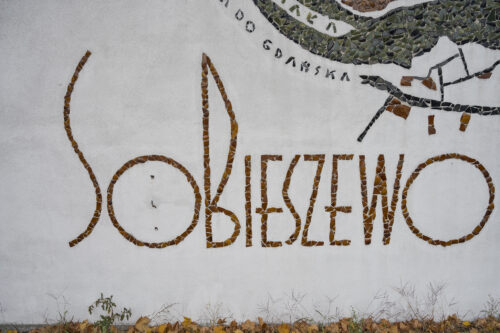 Zdjęcie poziome ze zbliżeniem na fragment wielokolorowej mozaiki z napisem: Sobieszewo, która pokrywa boczną ścianę budynku gospodarczego.