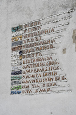 Zdjęcie poziome ze zbliżeniem na fragment wielokolorowej mozaiki z napisami ułożonymi pionowo obok kamieni w różnych kolorach. Ich układ przypomina swoim wyglądem legendę mapy lub planu.