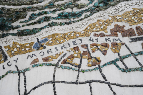 Zdjęcie poziome ze zbliżeniem na fragment wielokolorowej mozaiki. Przedstawia ona plan urbanistyczny a także linię brzegową.