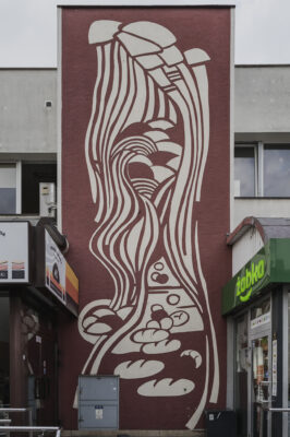 Zdjęcie pionowe przedstawiające boczną ścianę budynku – pawilonu handlowego, która w całości została pokryta dekoracją w technice sgraffito. Utrzymana jest ona w bordowo-kremowej kolorystyce i przedstawia m.in. bochenki chleba i rogale, a także różne formy geometryczne.