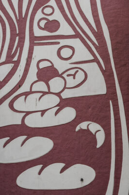 Zdjęcie pionowe ze zbliżeniem na fragment dekoracji bocznej ściany budynku – pawilonu handlowego, Została ona wykonana w technice sgraffito i jest utrzymana w bordowo-kremowej kolorystyce. Dekoracja przedstawia m.in. bochenki chleba i rogale, a także różne formy geometryczne.