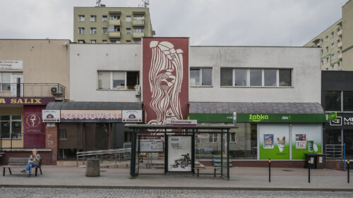 Zdjęcie poziome przedstawiające boczną ścianę budynku – pawilonu handlowego, która w całości została pokryta dekoracją w technice sgraffito. Utrzymana jest ona w bordowo-kremowej kolorystyce i przedstawia m.in. bochenki chleba i rogale, a także różne formy geometryczne.