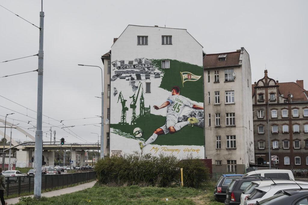 Zdjęcie ukazuje mural na fasadzie bocznej kamienicy. Mural przedstawia piłkarza podczas gry w piłkę w realistycznej stylistyce, powyżej postaci widoczny jest napis Lechia Gdańsk.
