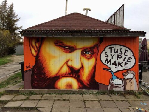 Zdjęcie ukazuje niski budynek, którego cała boczna ściana wypełniona jest graffiti. Na czerwonym tle widoczna jest duża twarz brodatego mężczyzny namalowana realistycznie, obok pole z napisem tuse mąke sypie, poniżej dwa worki z mąką.