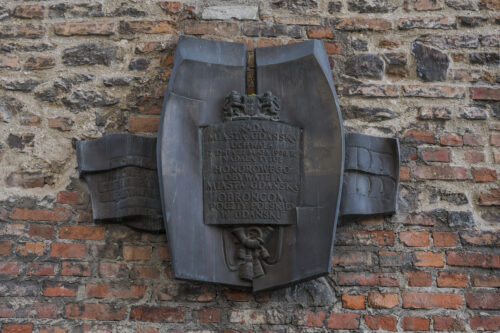 Zdjęcie ukazuje formę z brązu na której umieszczono napisy, jest to fragment rzeźby wkomponowanej w ścianę murowaną z cegieł.