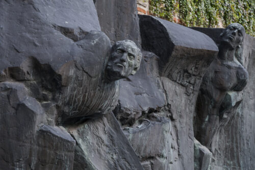 Zdjęcie ukazuje rzeźbę z brązu z patyną, na której można zauważyć postacie ludzkie wyłaniające się z form przypominających mur lub gruzy. Rzeźba została wkomponowana w ścianę z cegieł.