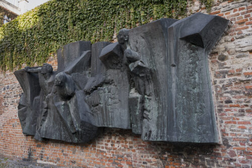 Zdjęcie ukazuje rzeźbę z brązu z patyną, na której można zauważyć postacie ludzkie wyłaniające się z form przypominających mur lub gruzy. Rzeźba została wkomponowana w ścianę z cegieł.
